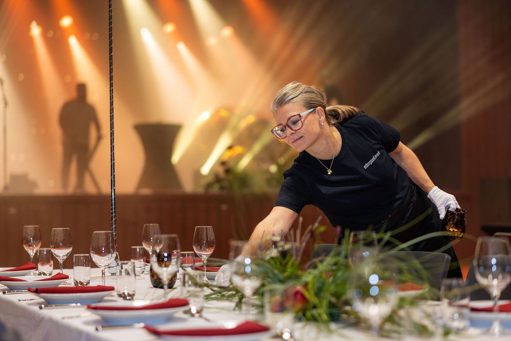 A catering staff member sets an elegant banquet table with wine glasses and red napkins before a stage event.