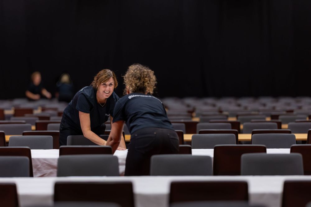 Two women in black uniforms arranging chairs in a large auditorium or theater venue.