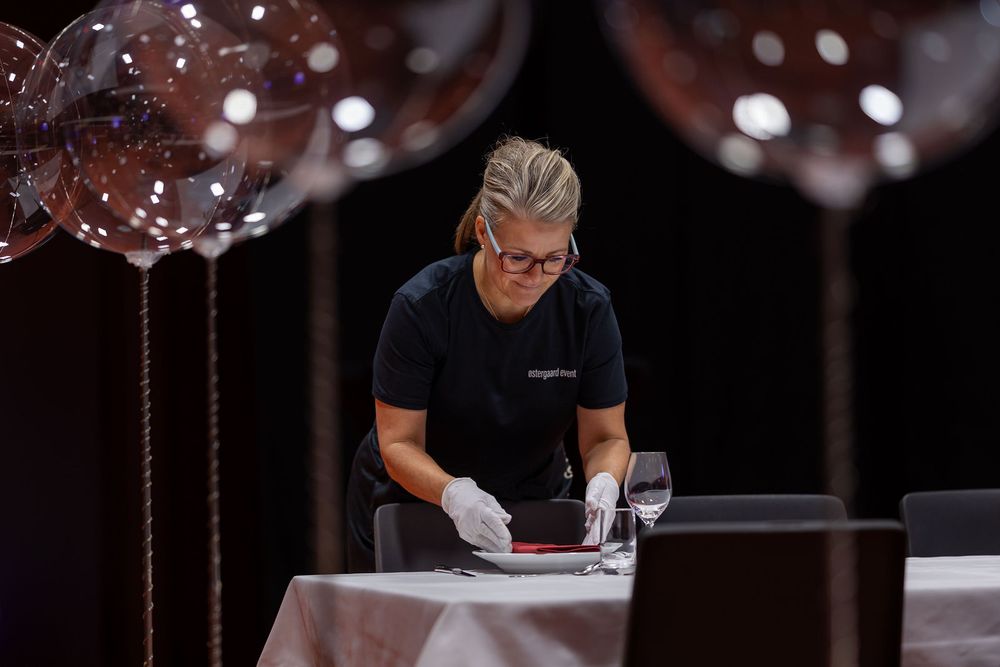 A woman in a black uniform carefully sets a table with white linens, surrounded by decorative balloons at an event.