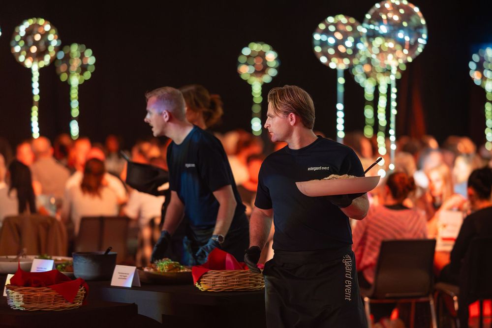 Catering staff serve guests at an elegant evening gala event with glowing floral light installations in the background.