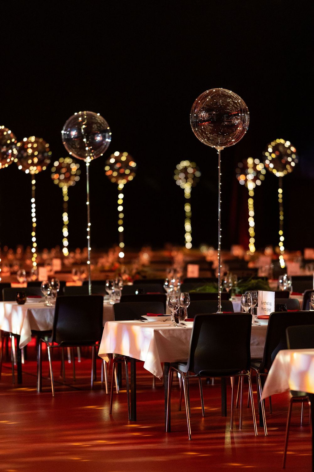 Elegant event venue with white-clothed tables, black chairs, and sparkling balloon centerpieces glowing against a dark night backdrop.