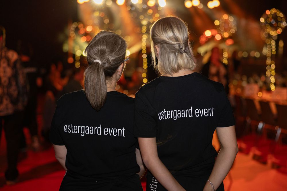 Two women in black Ostergaard Event t-shirts stand together at a colorful evening event.
