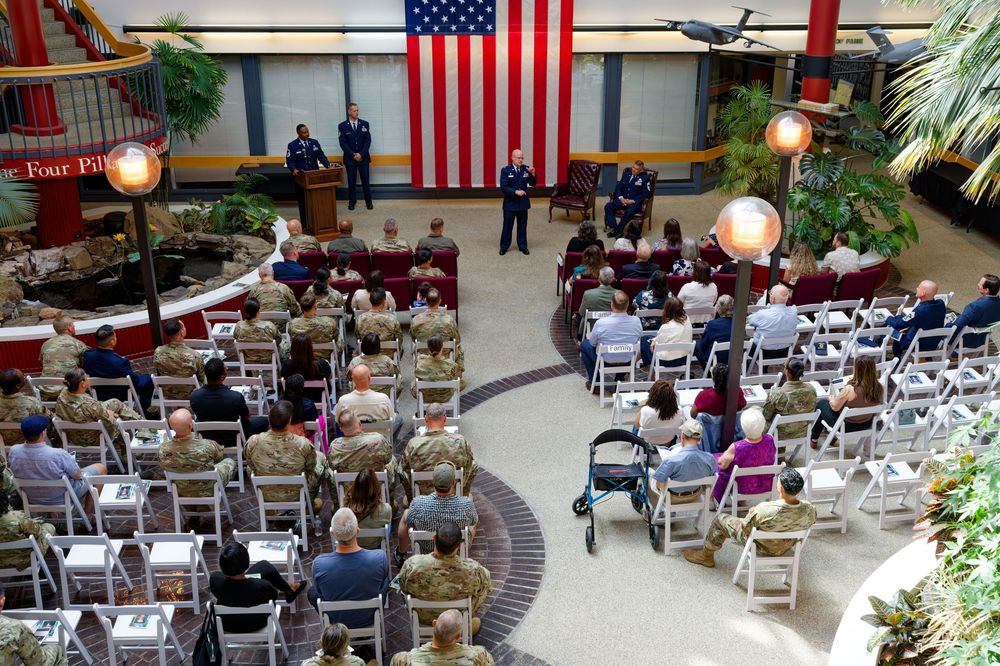 Military retirement ceremony in atrium with uniformed personnel, American flag, and seated audience.