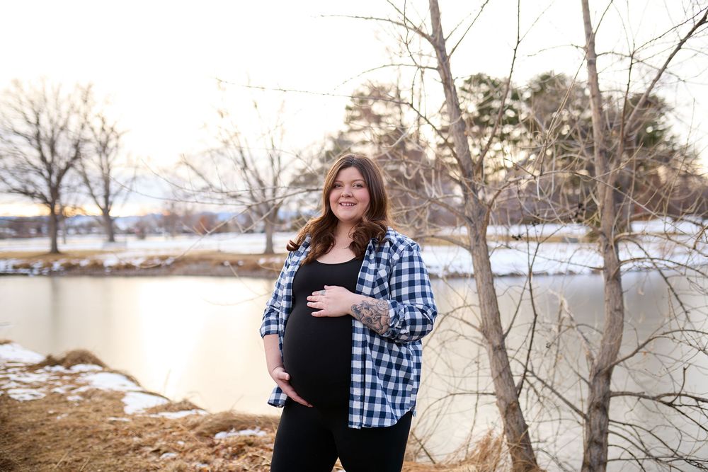 Pregnant woman in a plaid shirt cradles her belly while standing by a calm lake with bare winter trees and snow-dusted shoreline.