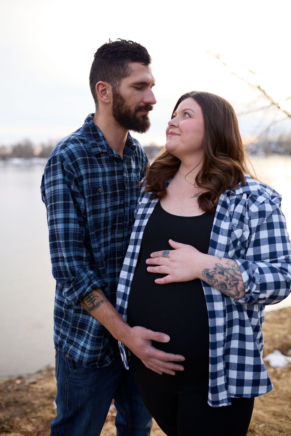 Bearded man and pregnant woman in plaid shirts share a tender moment by a lake during an outdoor maternity photo session.