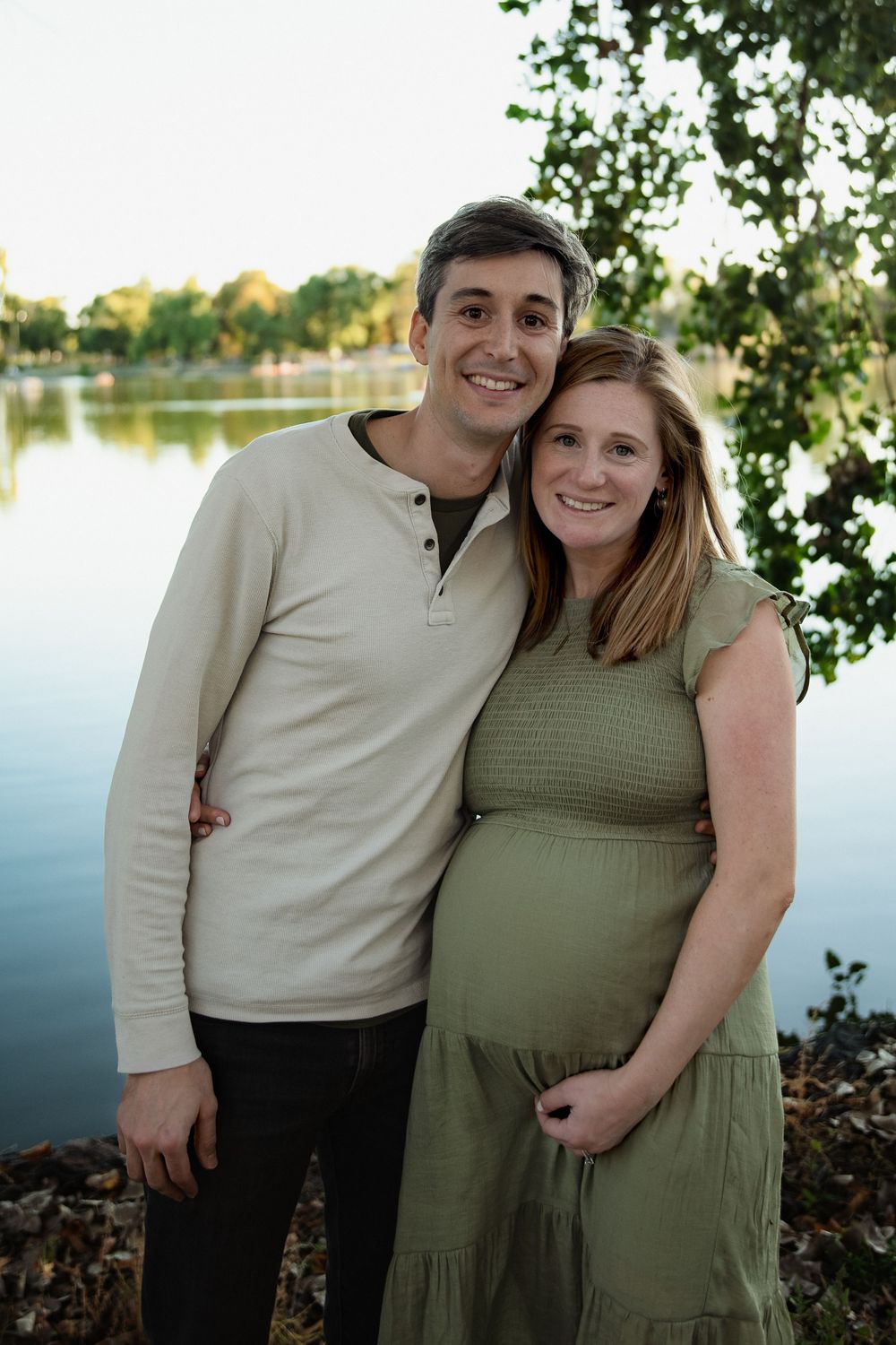 Smiling couple embracing outdoors by a calm lake at sunset, the pregnant woman in a green dress cradling her baby bump beside her partner.
