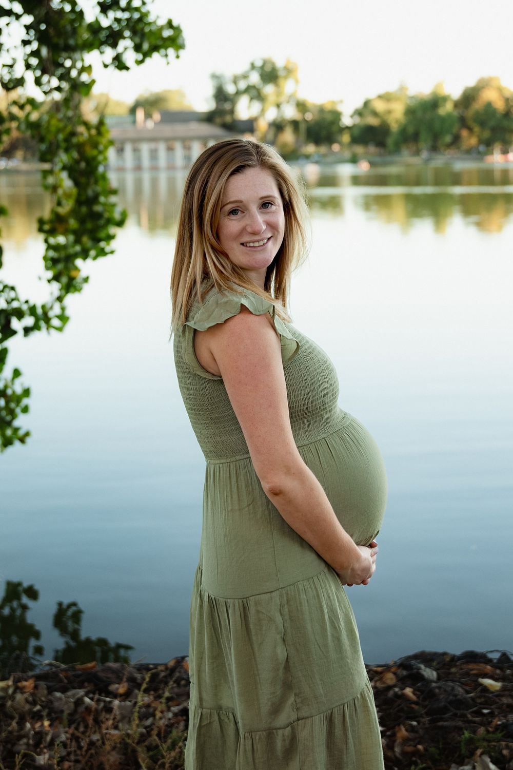 Smiling pregnant woman in a green dress cradles her baby bump while standing beside a calm lake with trees in the background.
