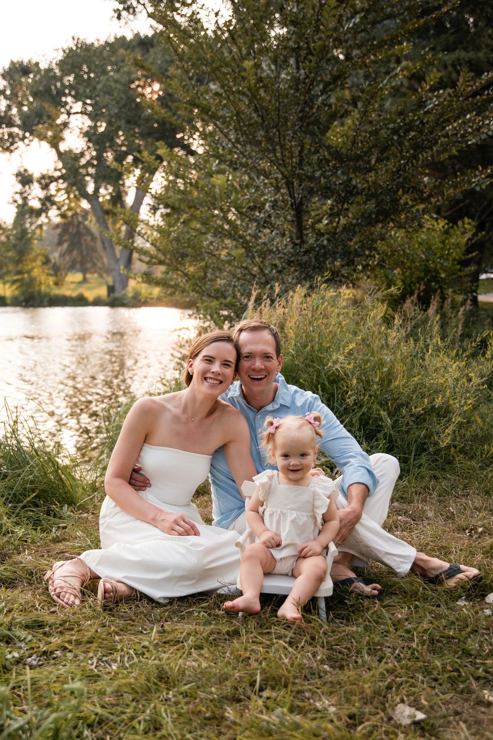 A smiling couple in white and blue sit on the grass with their toddler daughter beside a calm lake at sunset.