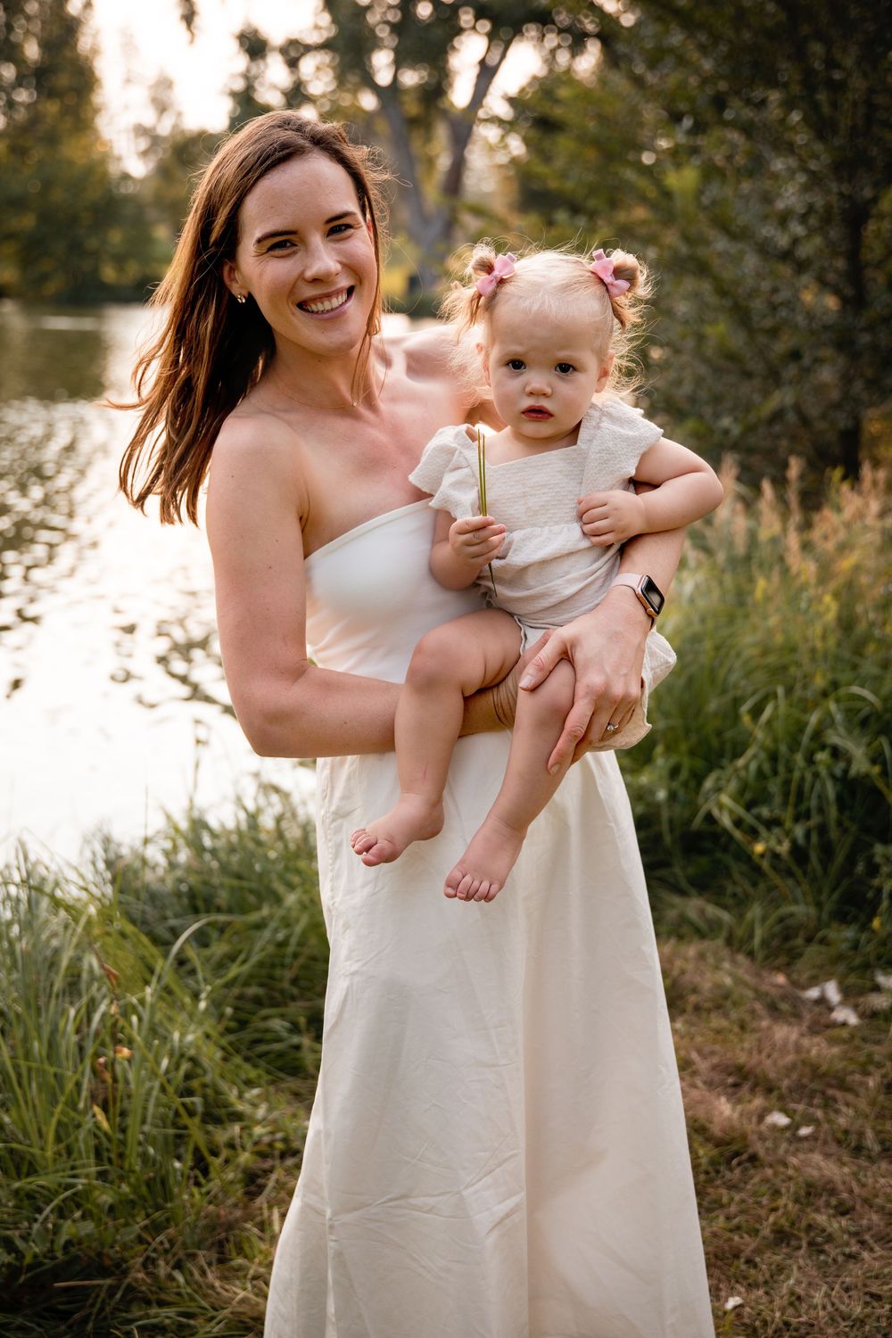 Smiling woman in a white dress holds a toddler with pink hair bows while standing in tall grass beside a lake at golden hour.