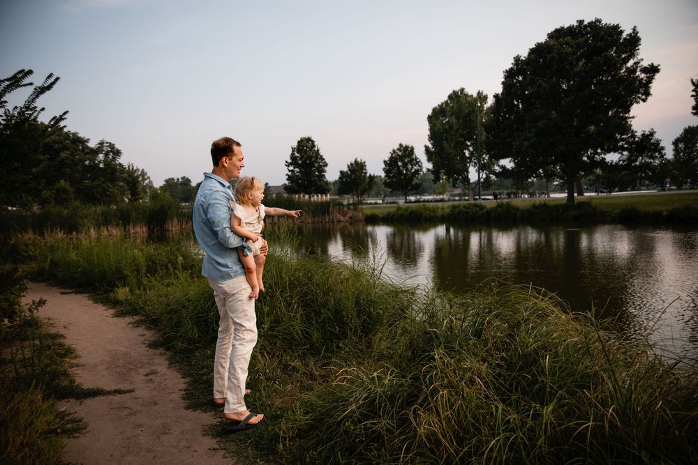 A father in a light blue shirt holds his young child while standing beside a calm pond with tall grass and trees at sunset.