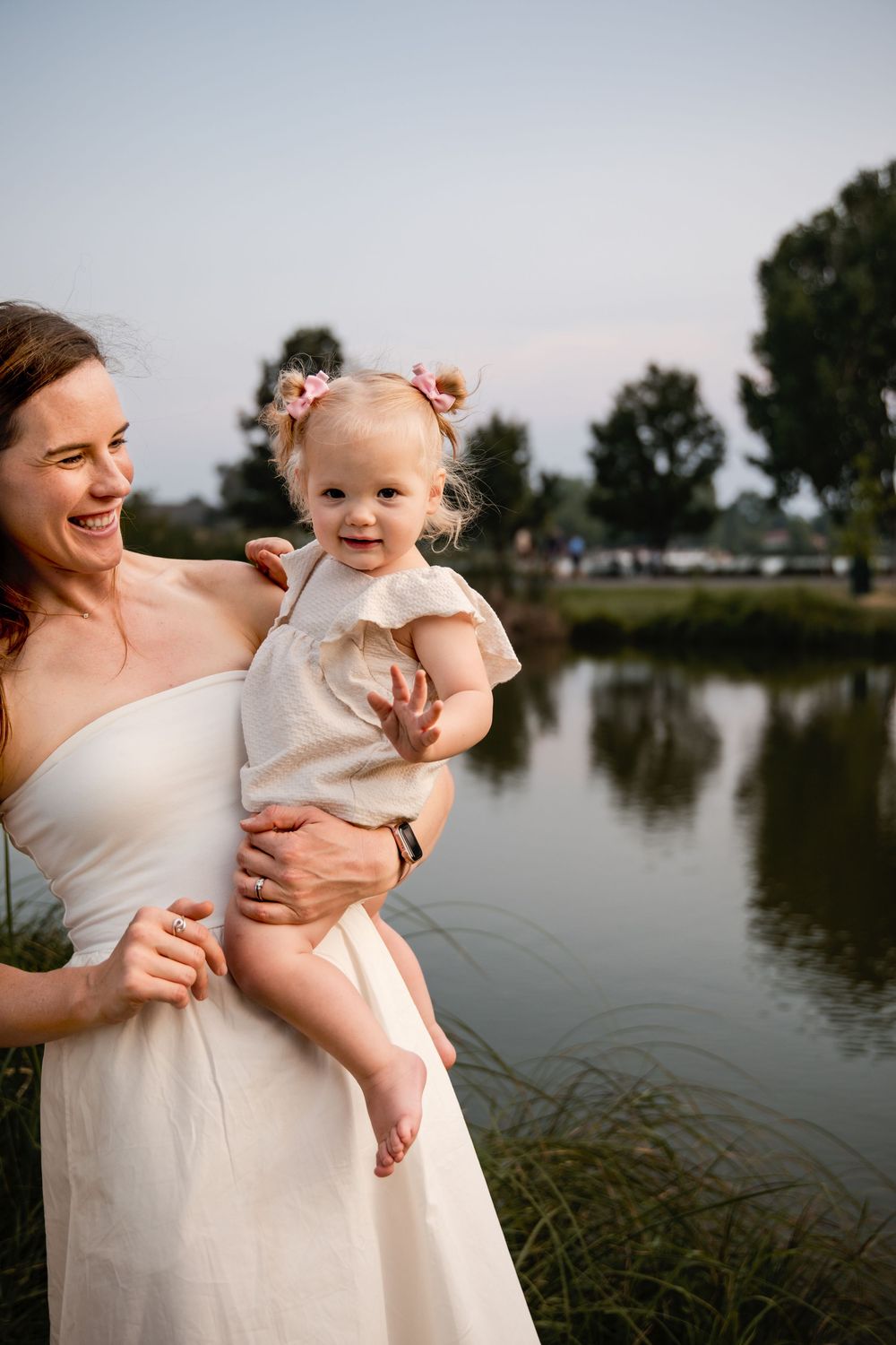 Smiling woman in a cream dress holding a toddler with pink hair bows beside a calm lake at sunset, with trees reflected in the water.