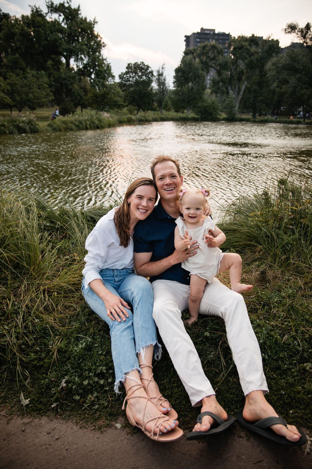 A smiling couple sits on the grassy bank of a sunlit pond holding their baby, with trees and a tall building in the background.