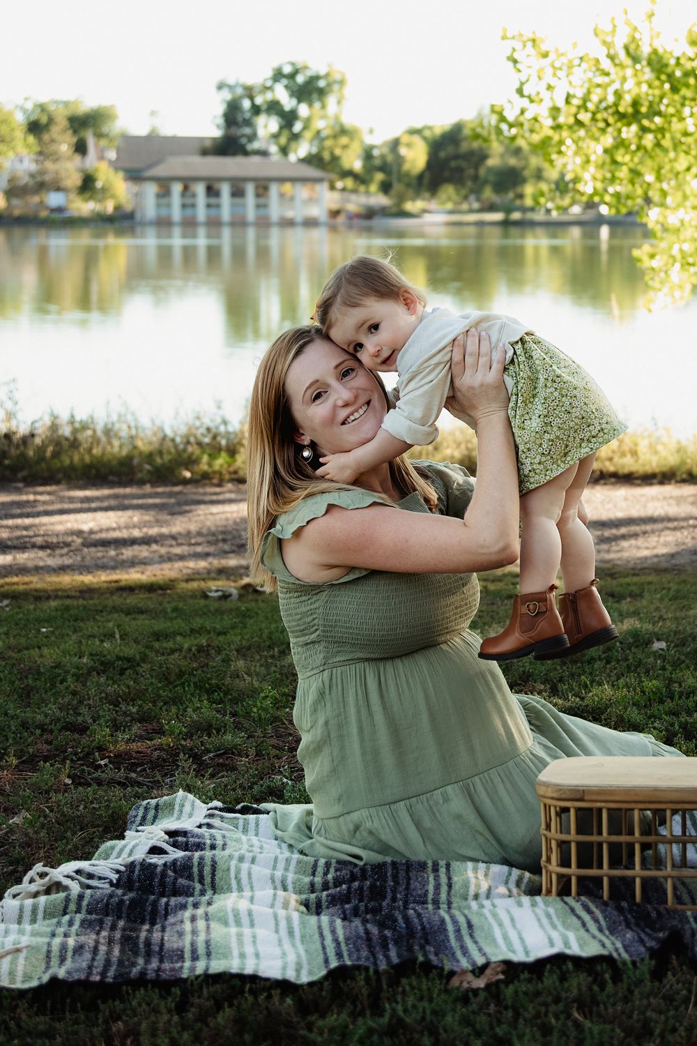 Smiling woman in a green dress sits on a plaid picnic blanket by a lake, lifting a laughing toddler in a floral skirt and brown boots.