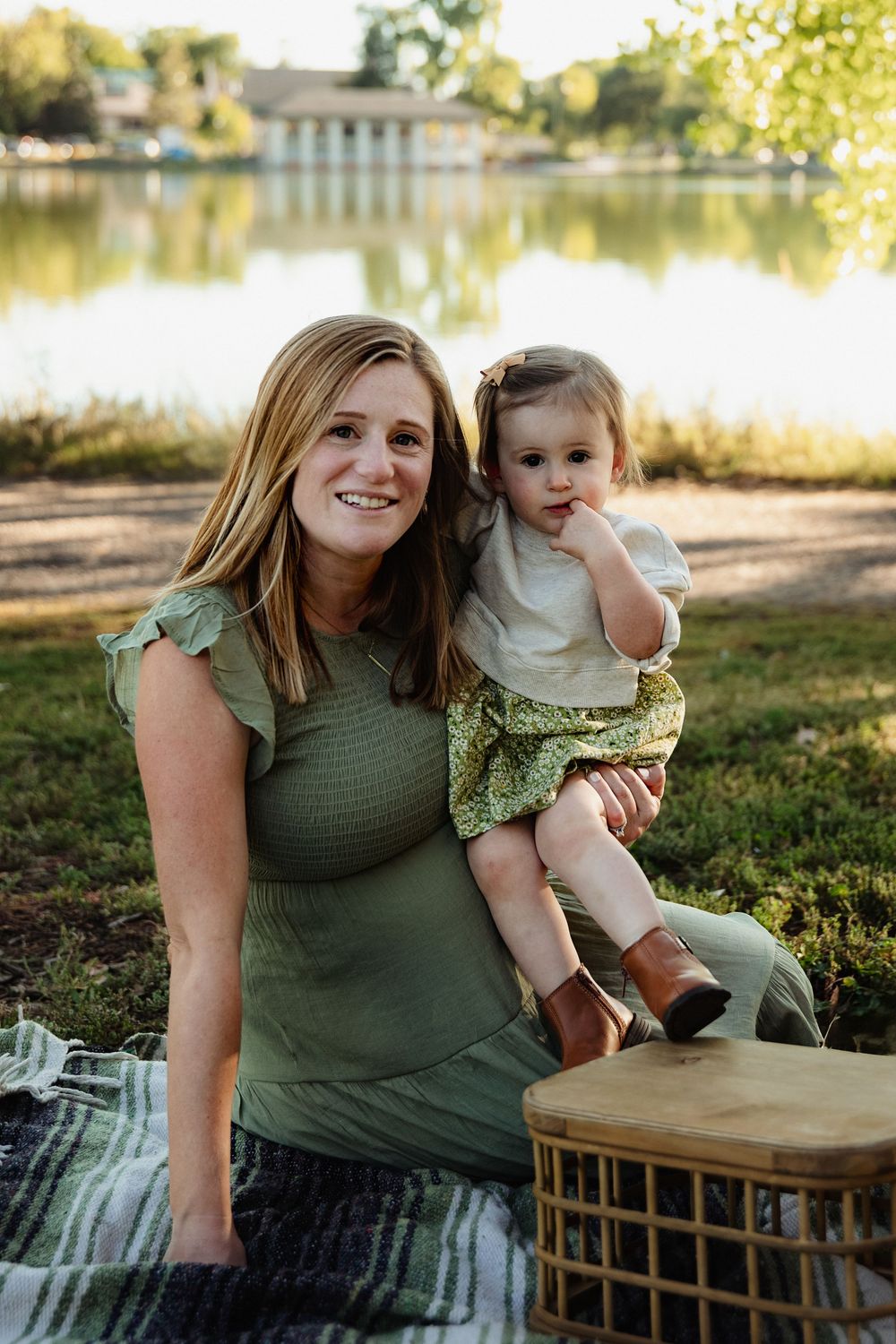 Smiling woman in a green dress sits on a picnic blanket holding a toddler in boots beside a wicker basket, with a calm lake behind them.