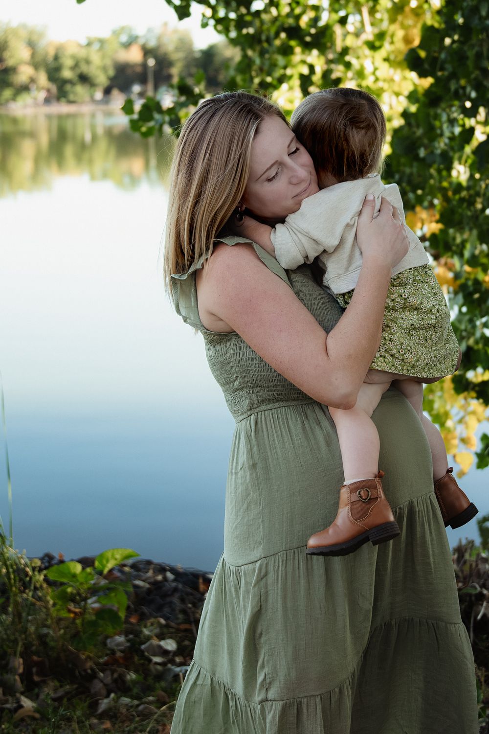 A woman in a green sundress holds a toddler in a matching dress and brown boots while standing beside a calm lake in autumn.