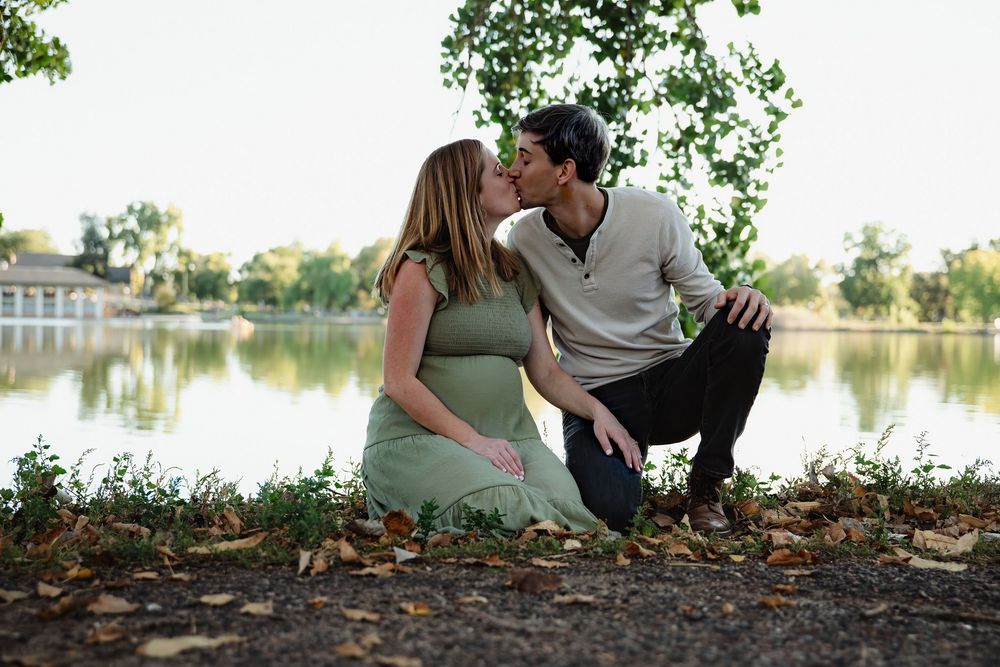 Expectant couple shares a kiss while sitting on the grass by a peaceful lake, framed by trees and autumn leaves in a maternity photo.