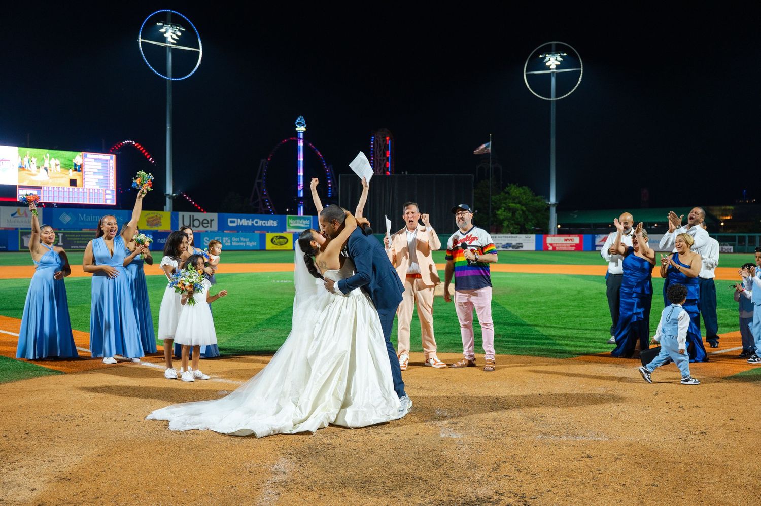 Bride and groom kiss on the Brooklyn Cyclones' baseball field at night while bridesmaids in blue dresses and groomsmen look on.
