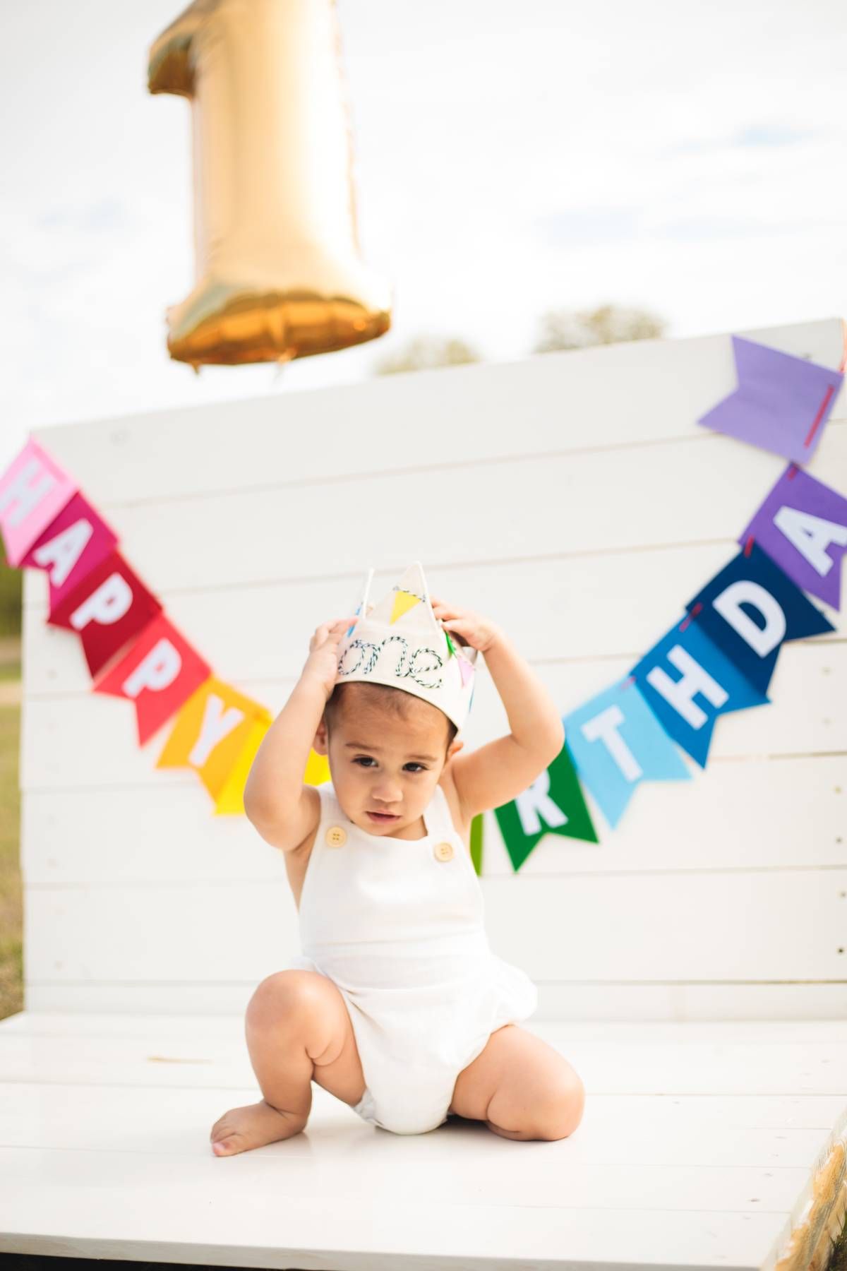 Baby in a white romper and paper crown sits on a white platform beneath a rainbow happy birthday banner and gold number one balloon.
