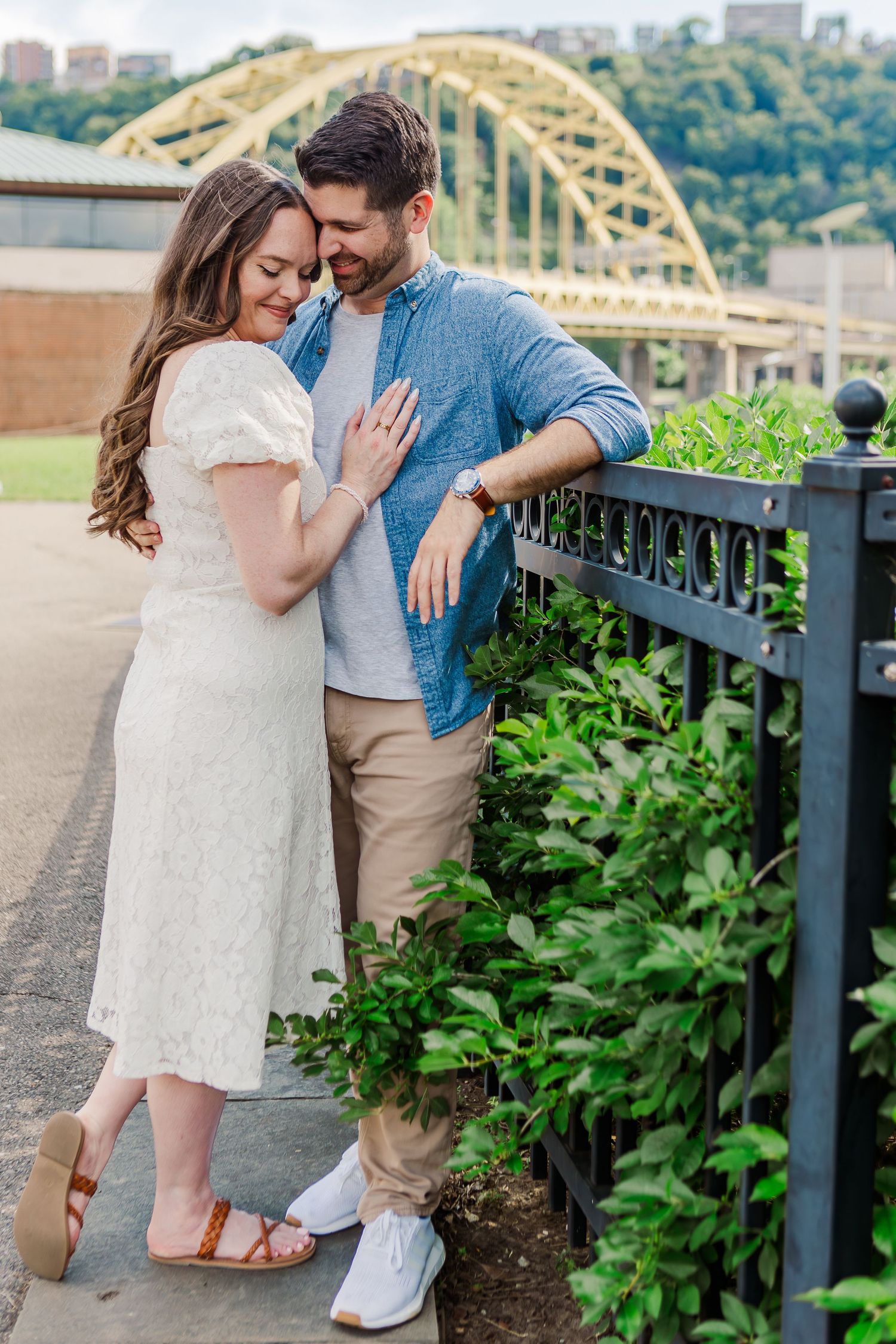 Couple embracing near black iron fence with Pittsburgh bridge and lush greenery in the background.