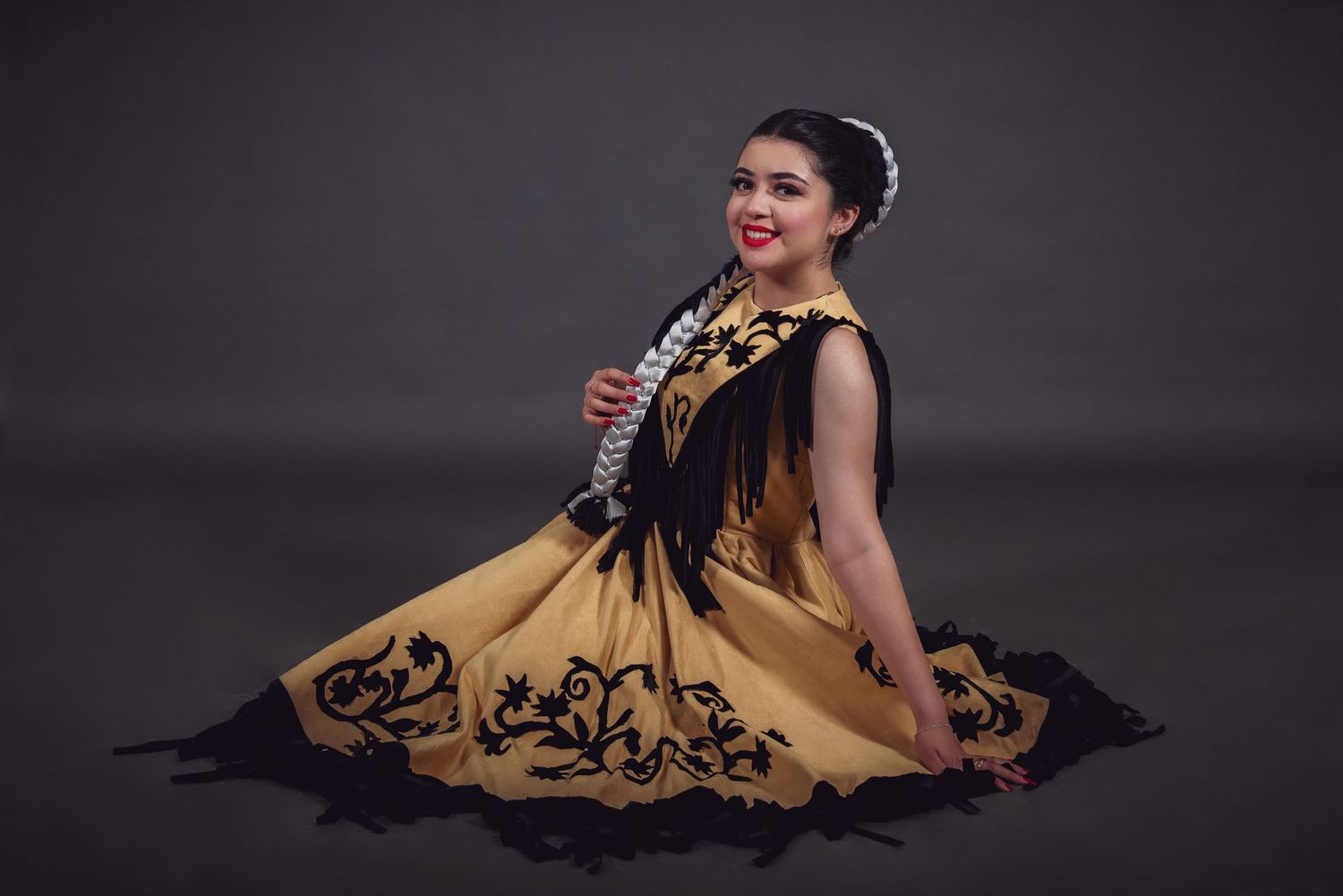 Smiling woman in a golden traditional dress with black embroidered trim sits gracefully on the floor against a dark studio backdrop.