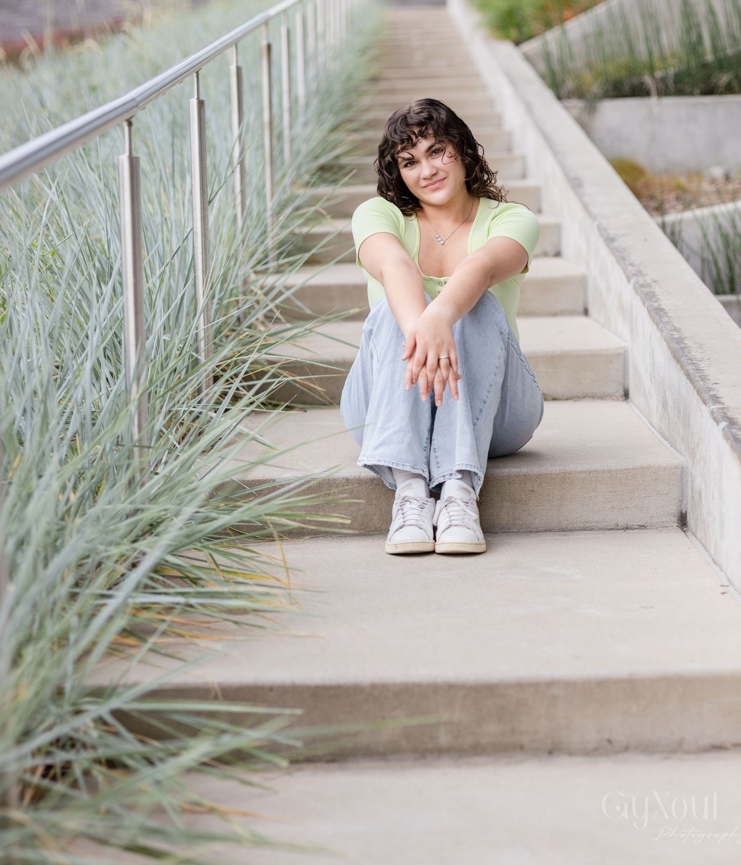 Young woman with curly hair sits on concrete steps, wearing a green top and jeans, smiling outdoors.