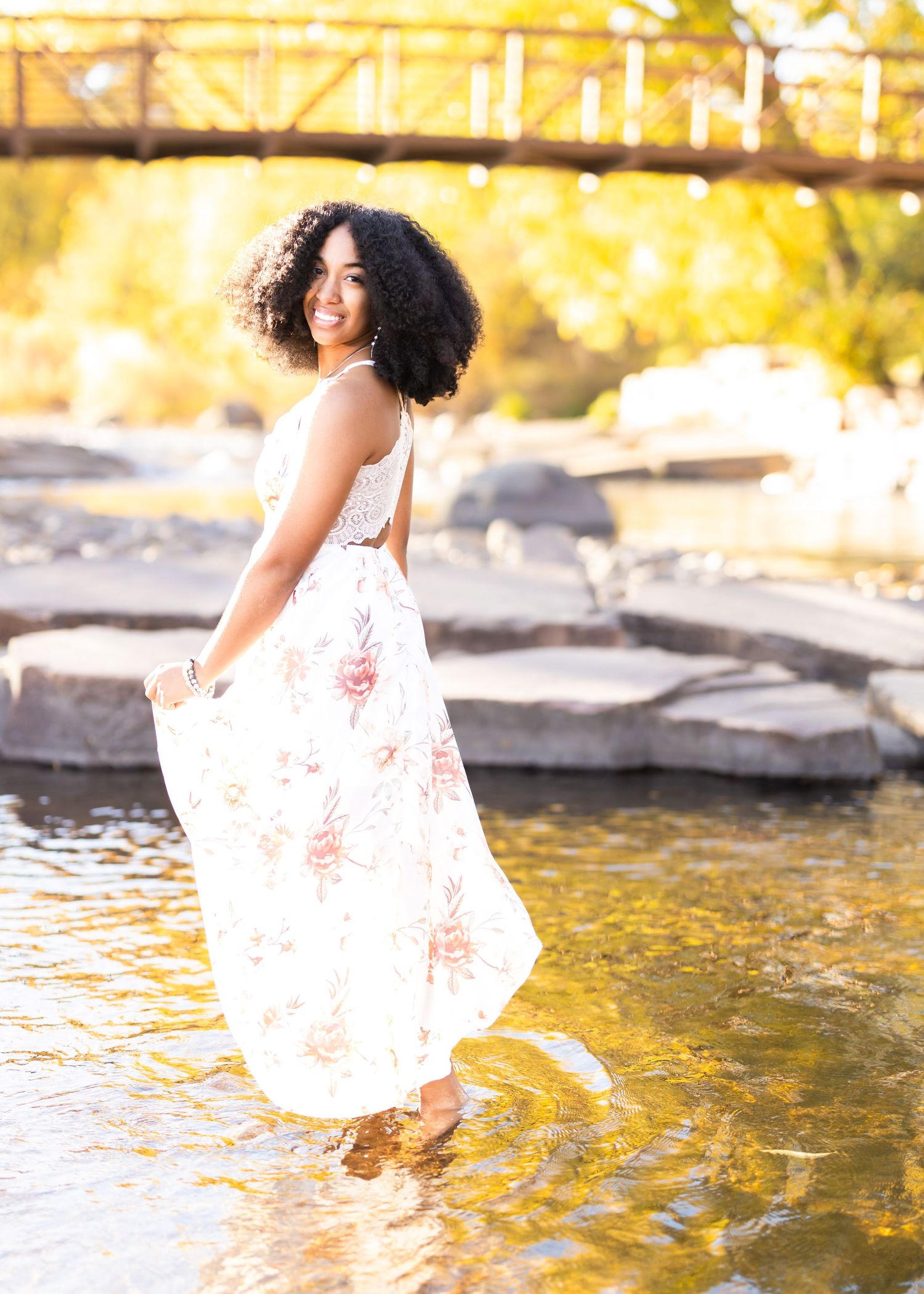 Young woman in floral maxi dress smiling by a rocky stream with a wooden bridge backdrop.