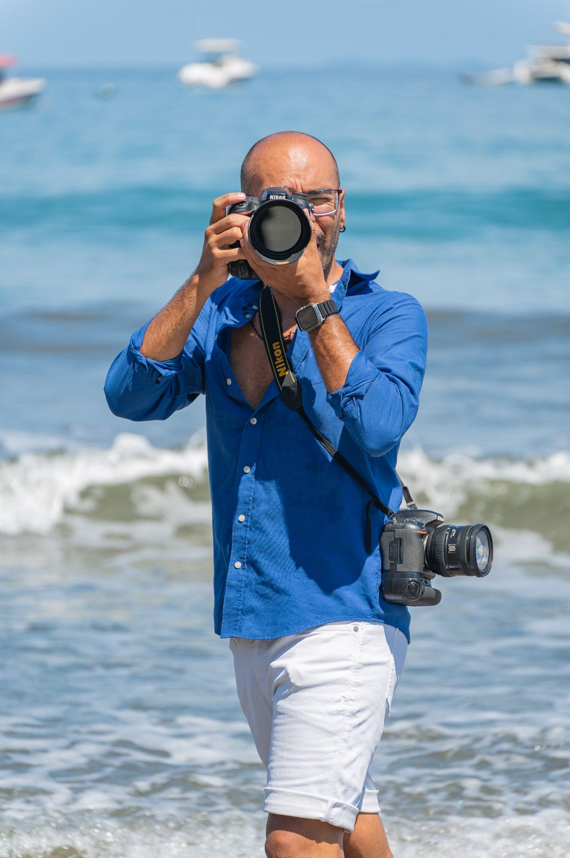 Bald man in blue shirt and white shorts photographing with Nikon DSLR camera at sunny beach with turquoise ocean waves.