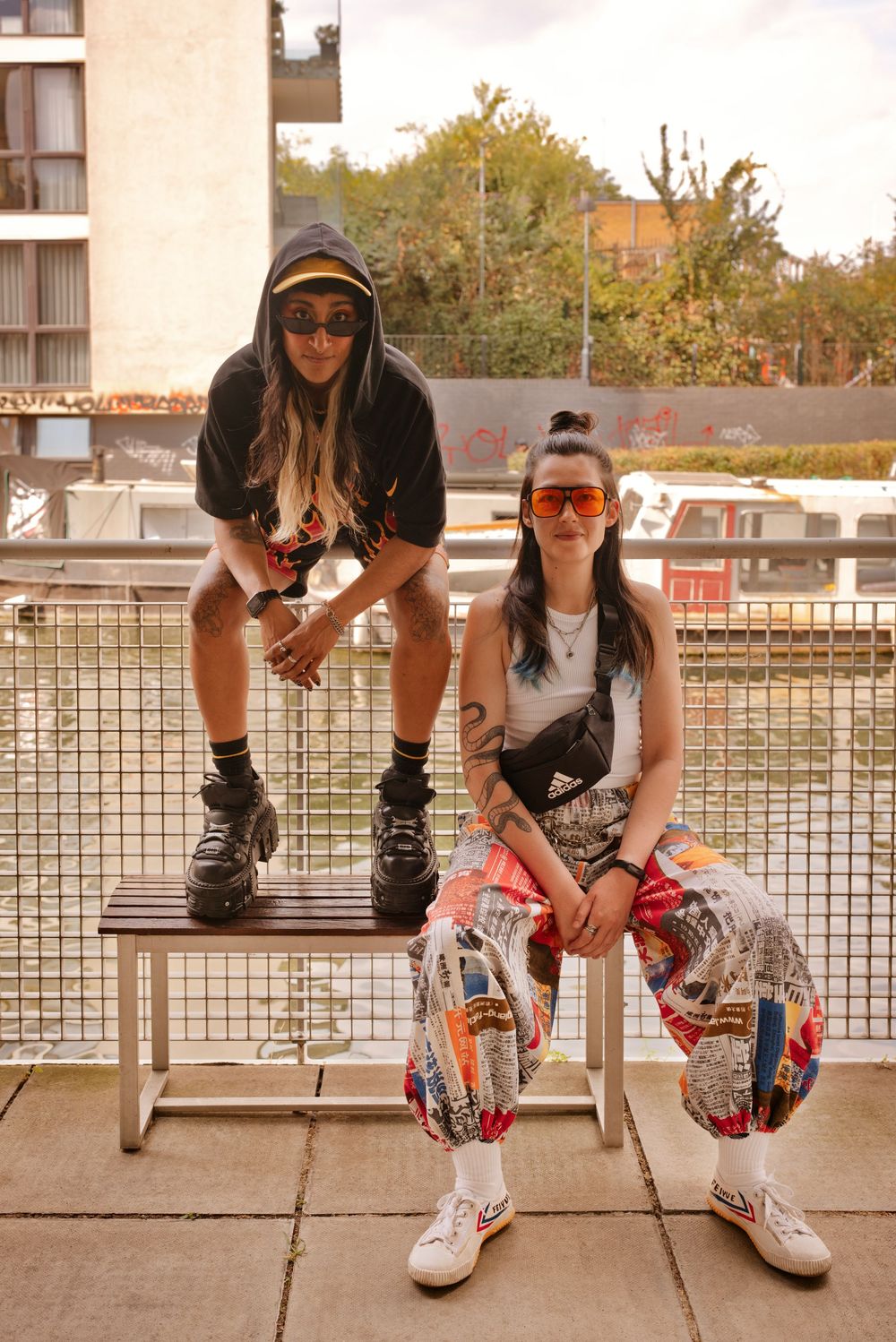 Two stylish women in colorful streetwear pose on an urban balcony, wearing bold sunglasses and trendy outfits.