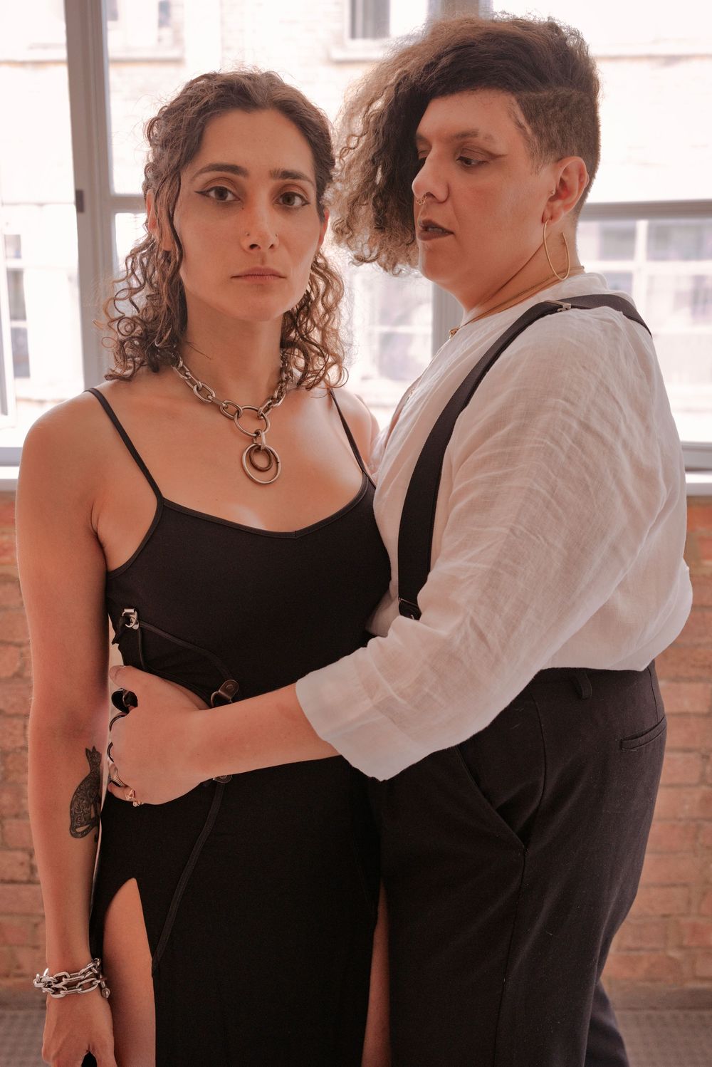 Two people posing together, one in a black dress with chain necklace, other in white shirt and suspenders.