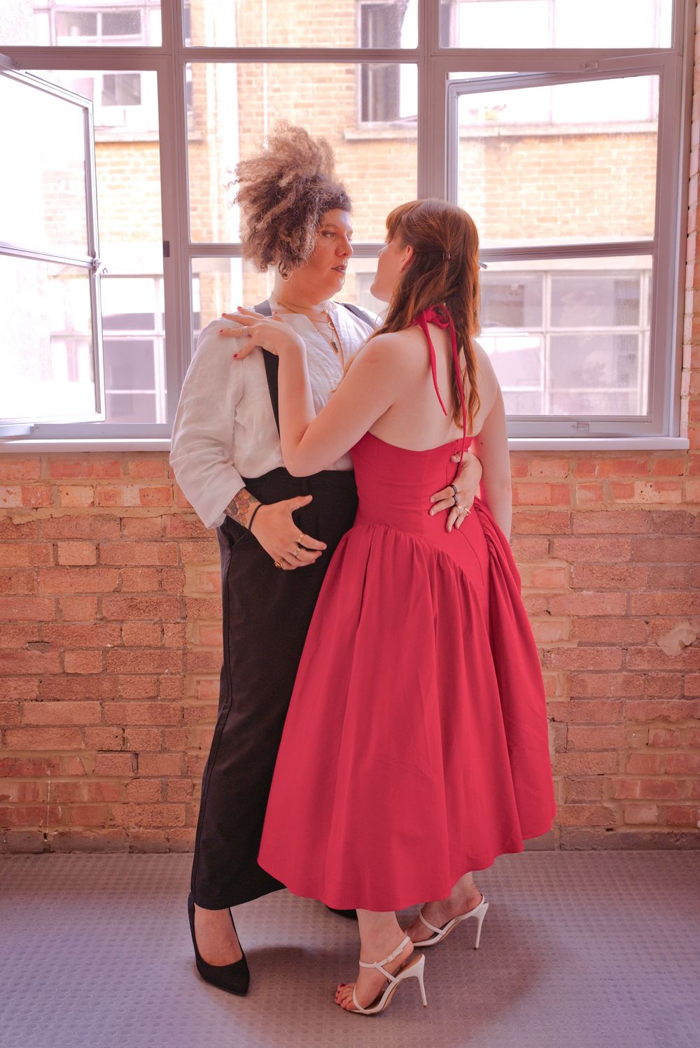 Two people in formal attire embrace near a brick wall, one in a red dress and one in black pants and white shirt.