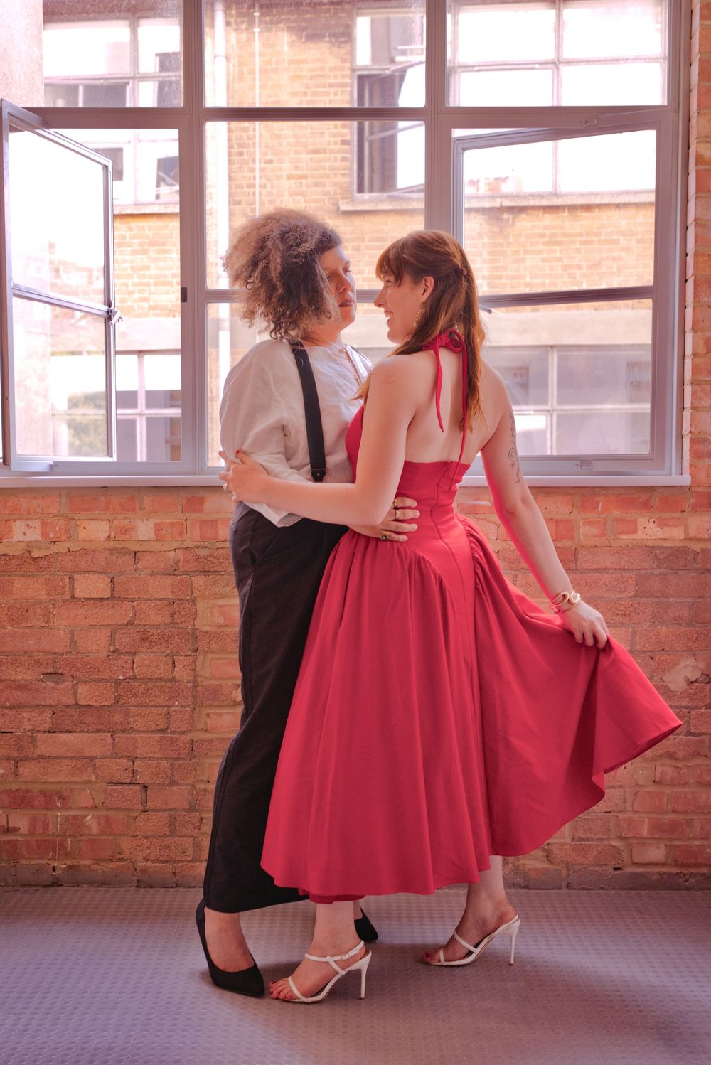 Two women in vintage attire dance together, one in a red halter dress, the other in suspenders, near a brick wall.