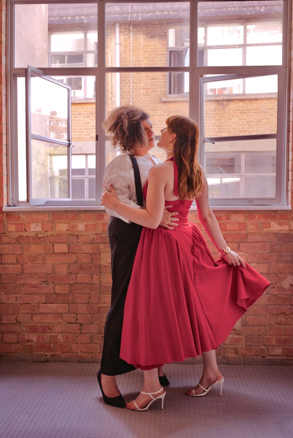 Couple in formal wear dancing romantically near brick wall window, woman in red dress, man in black trousers.