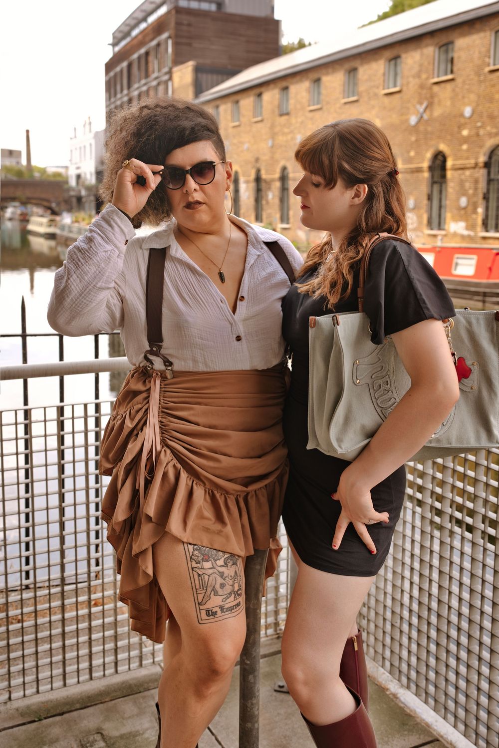 Two women posing on a bridge near a canal, one in a steampunk skirt with sunglasses, the other in a black dress with a tote bag.