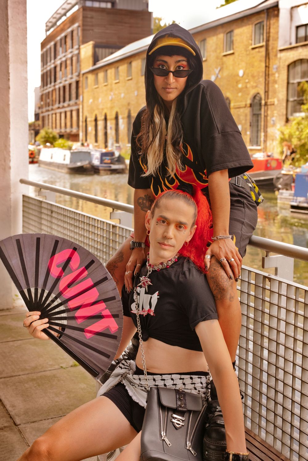 Two edgy fashion models pose by a canal in London, one holding a black and pink hand fan, wearing alternative streetwear.