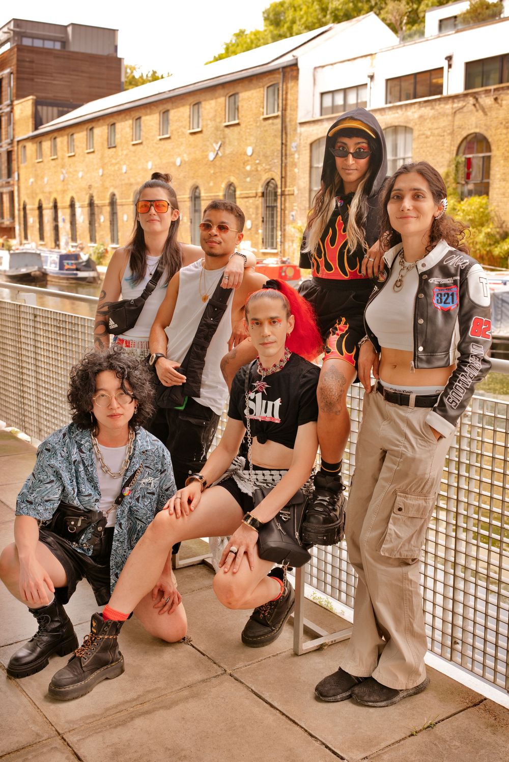 Six stylish young people posing outdoors near a canal in edgy streetwear fashion including flame prints and varsity jackets.