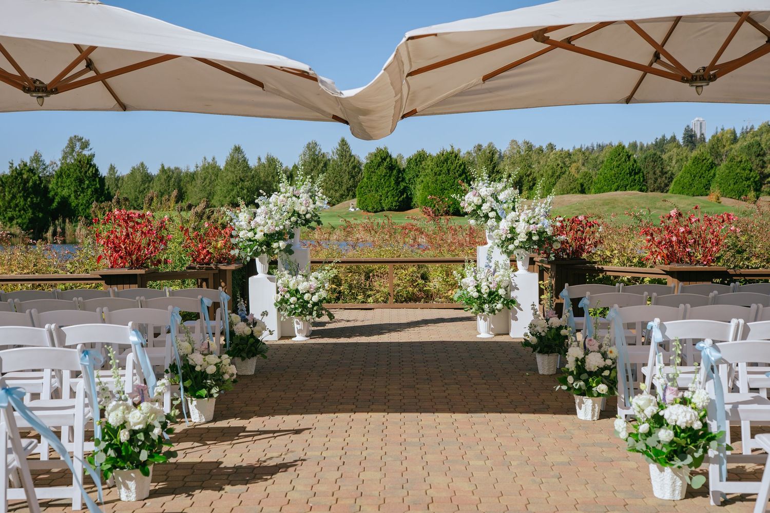 Elegant outdoor wedding ceremony aisle lined with white floral arrangements and chairs under large umbrellas.
