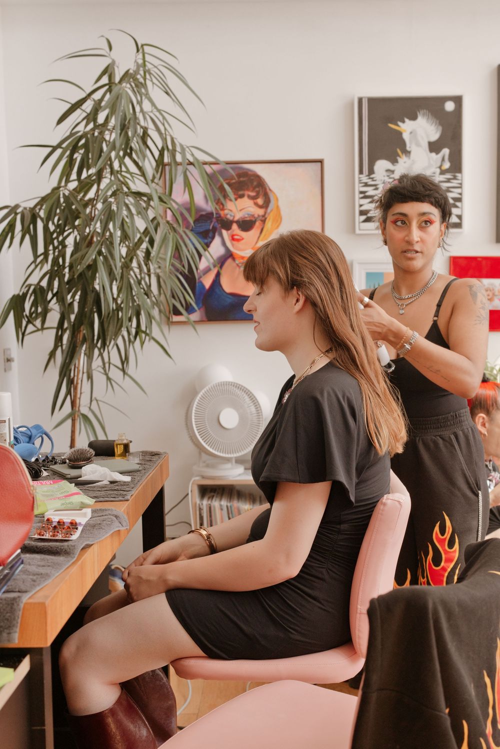 A hairstylist works on a seated woman in a black dress inside a vibrant studio with artwork and plants.