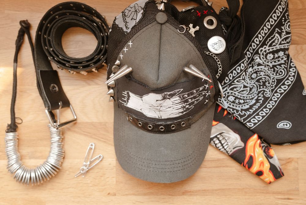 Gray studded baseball cap with bandana, leather belt, silver necklace, and flame accessories on wooden surface.