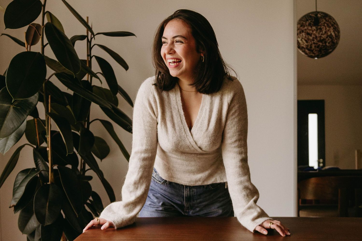 A young woman in a beige wrap sweater smiles warmly while leaning on a wooden table near a green plant.