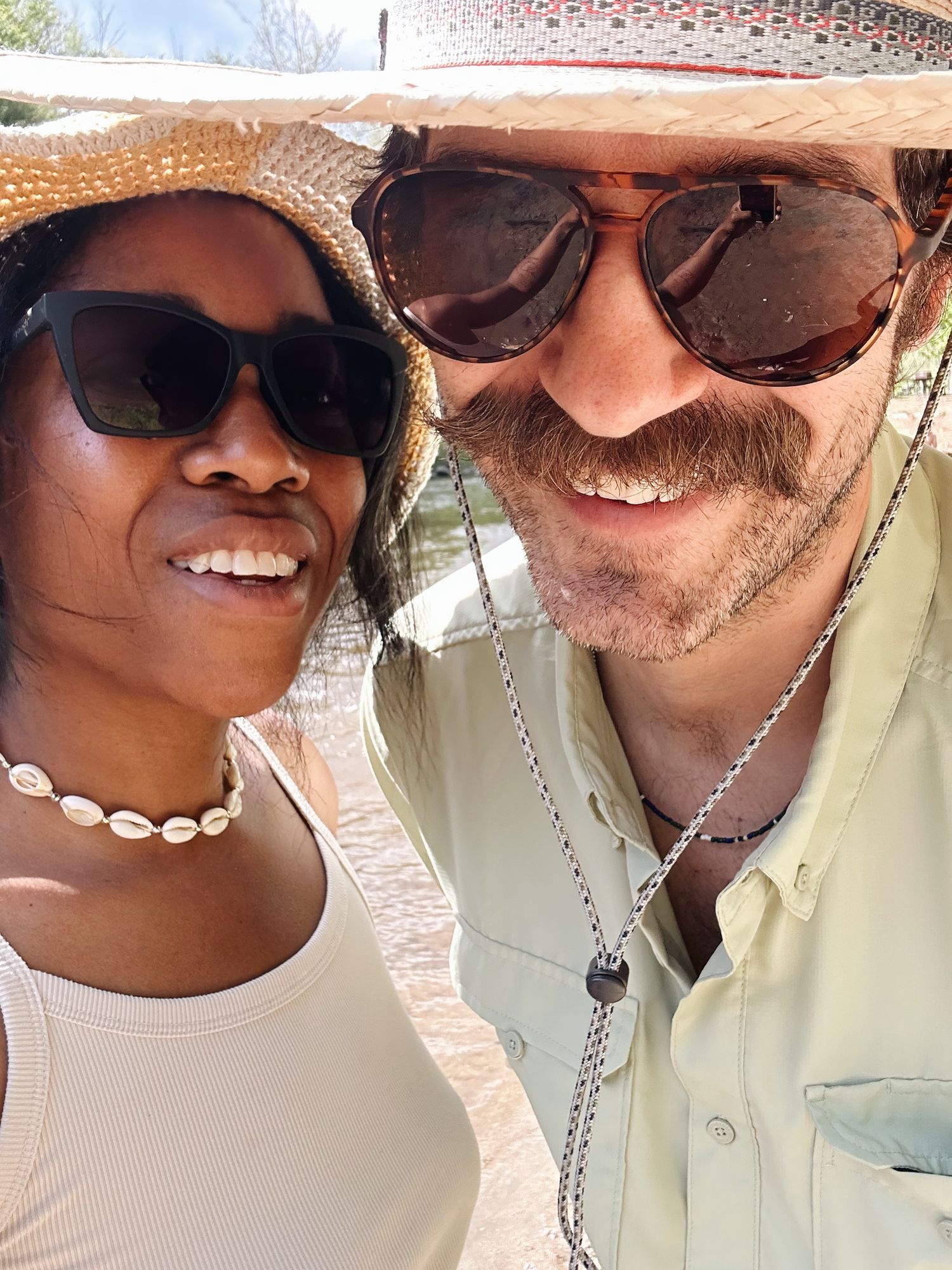Two smiling people wearing sunglasses and sun hats pose for a sunny outdoor selfie, woman wearing shell necklace.