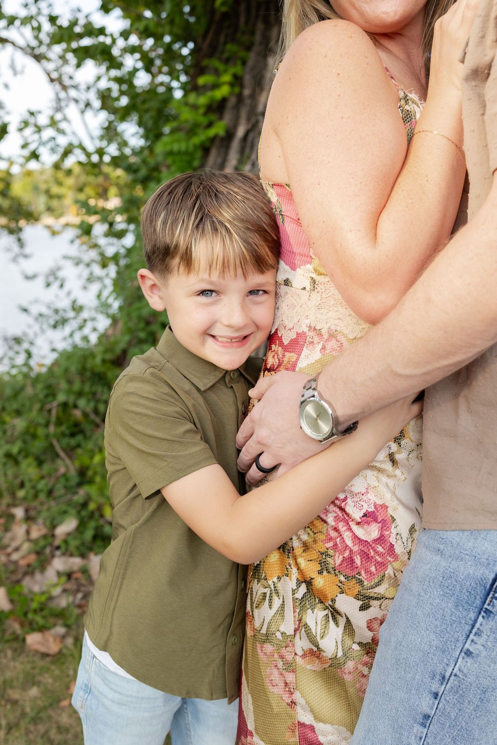 Smiling young boy in an olive green shirt hugging a woman in a pink floral sundress outdoors near a lake with trees.