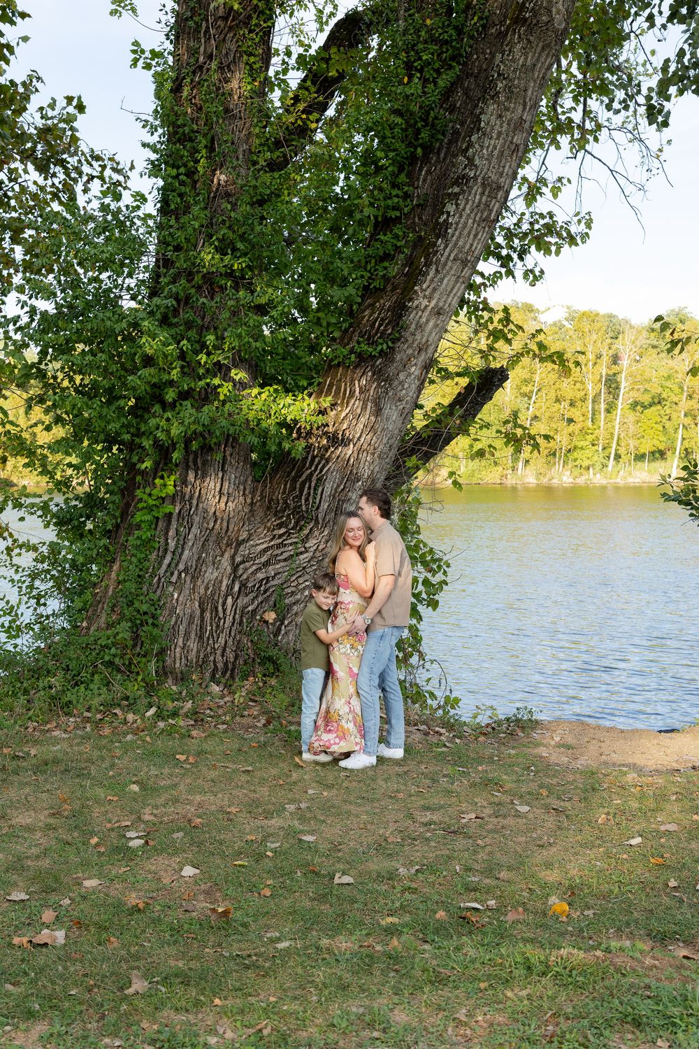 Family of three embracing beside a large ivy-covered tree on a grassy lakeside bank with autumn leaves and trees in the background.