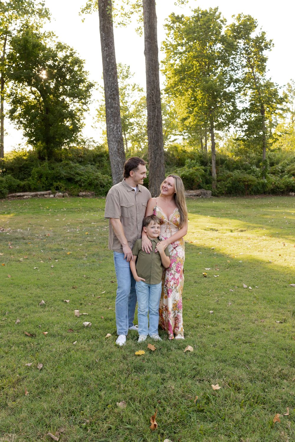 Smiling family of three poses together on a sunny grassy lawn with tall trees behind them during a golden hour outdoor portrait session.