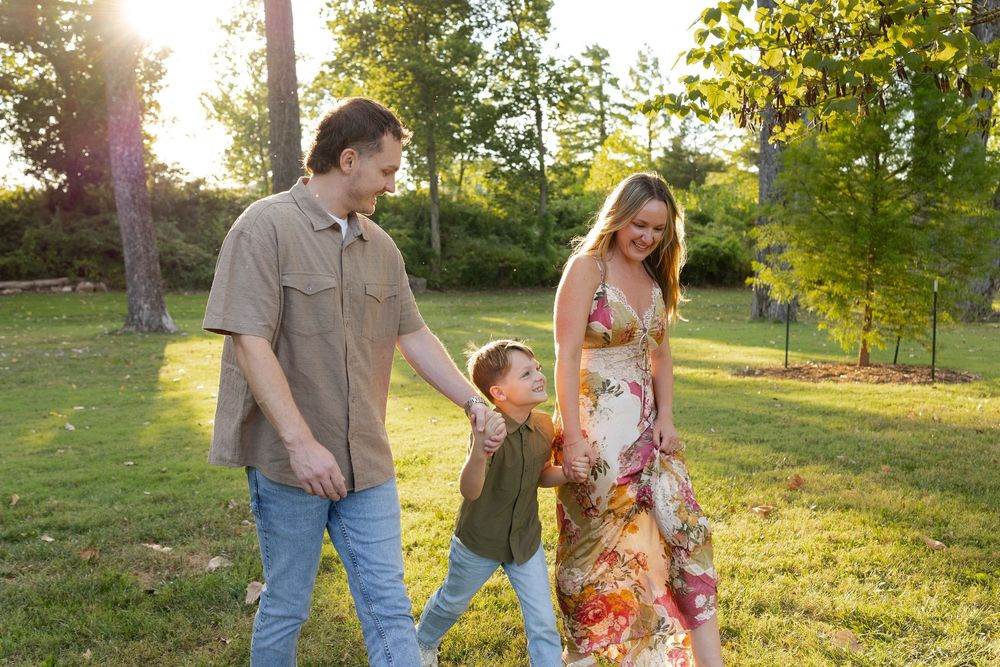 Family of three walks hand in hand across a sunlit grassy park, with a smiling woman in a floral dress, a man, and a young boy.