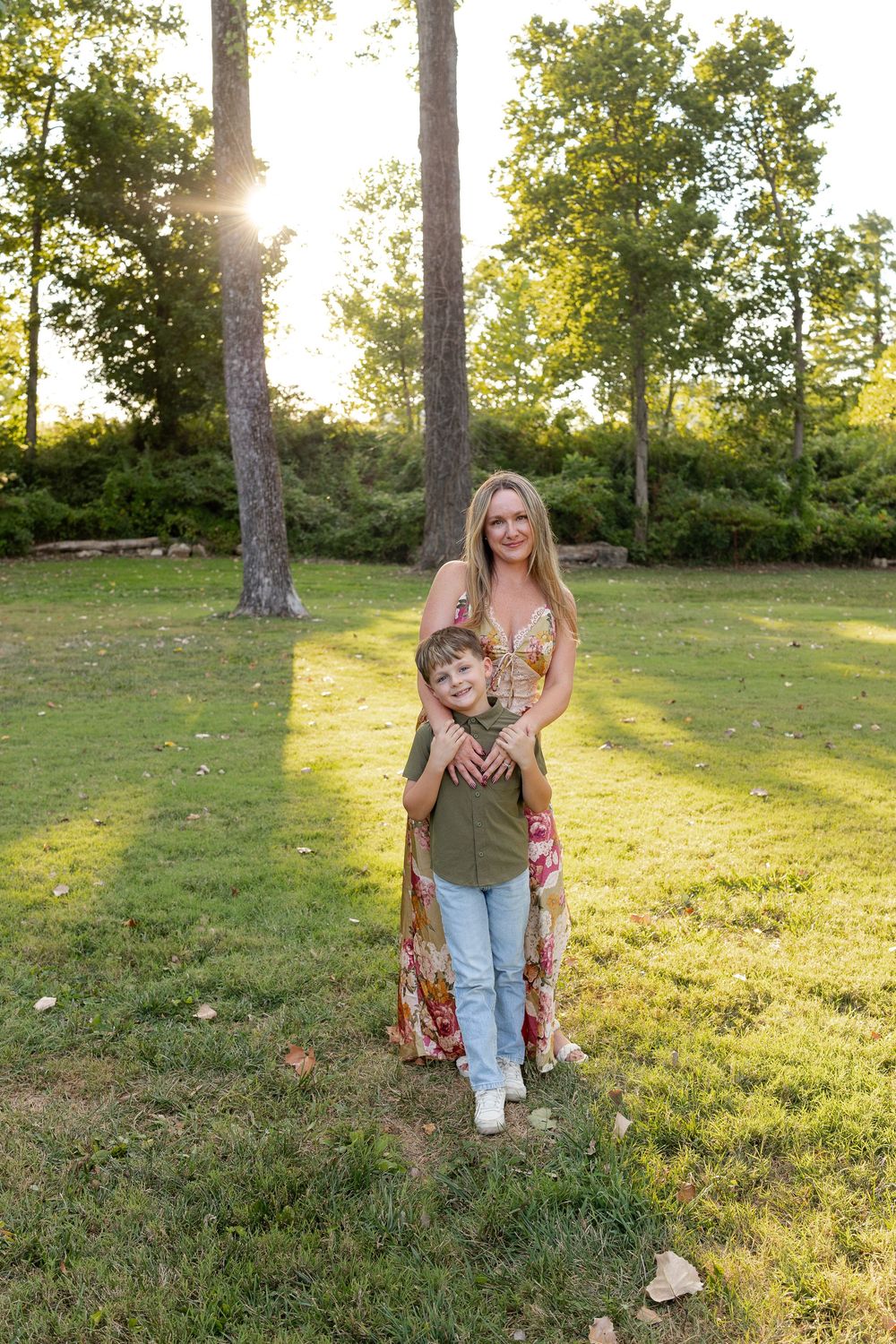 A smiling woman in a floral dress hugs a young boy from behind on a sunlit grassy lawn surrounded by tall trees.