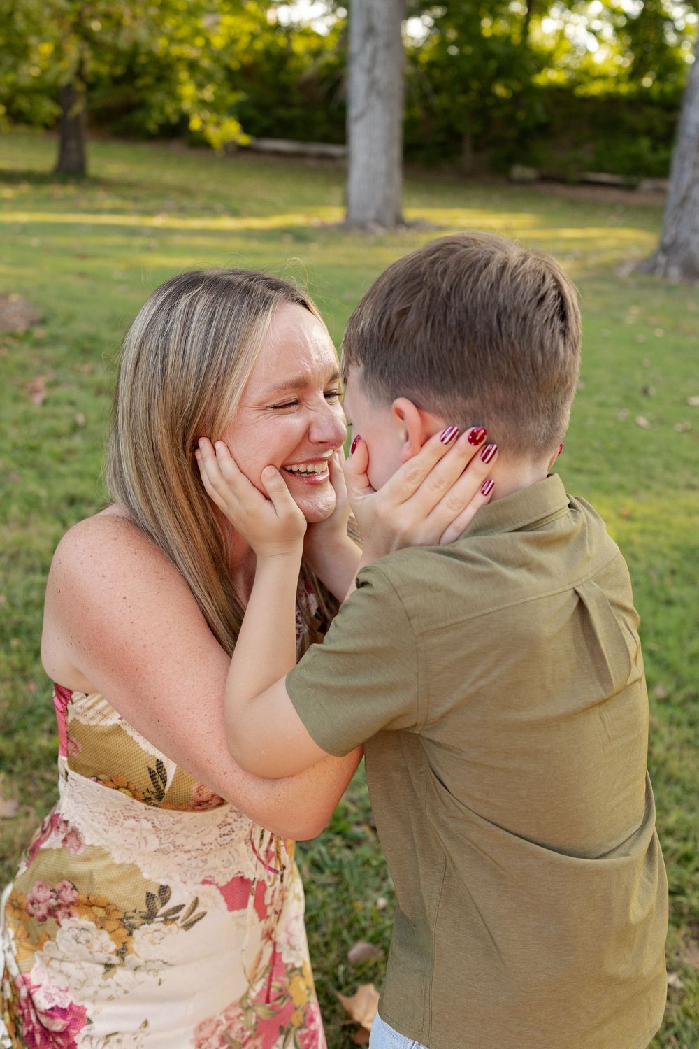 A smiling woman in a floral dress and a child in an olive shirt gently hold each other's faces in a sunny, tree-lined park.