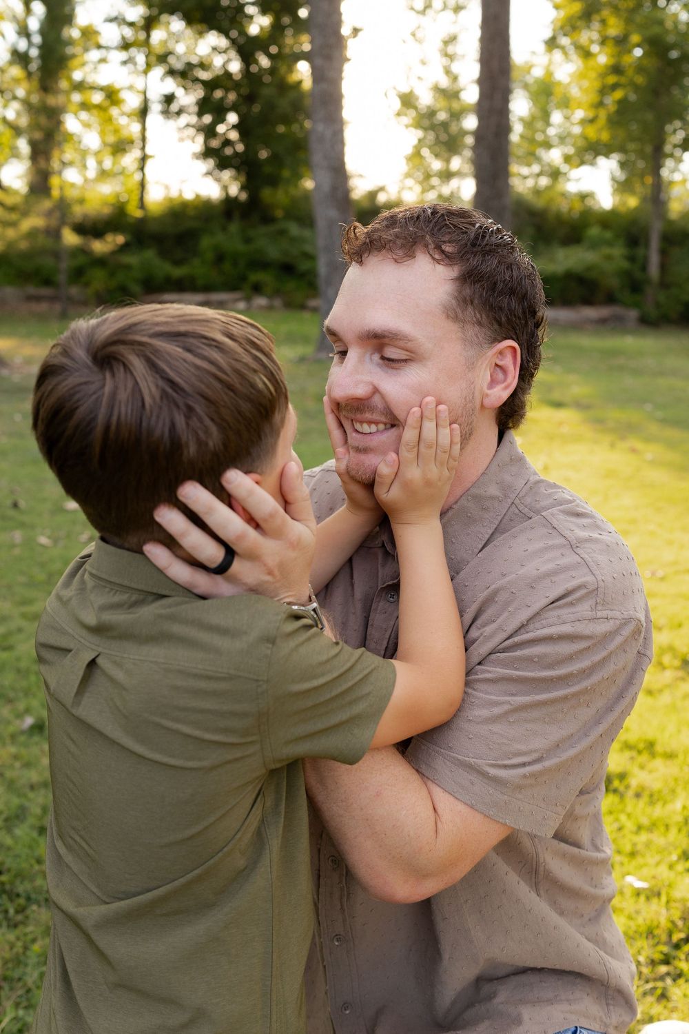A smiling man in a gray shirt embraces a young boy in a green shirt who gently holds his face in an outdoor park.