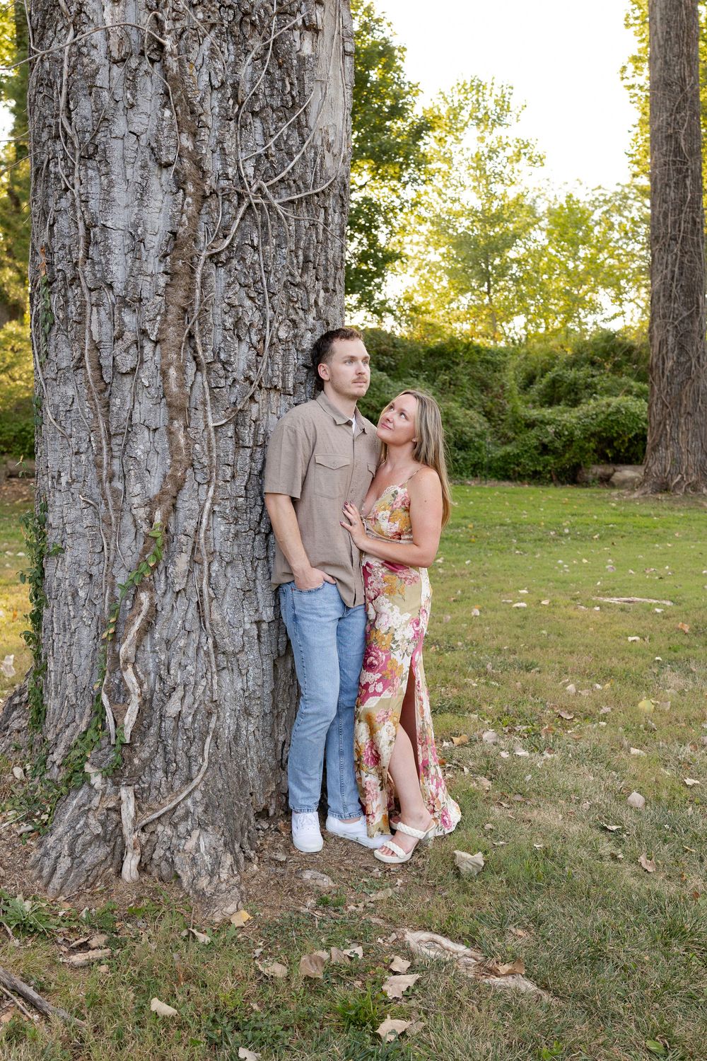 Couple posing against a large ivy-covered tree trunk in a sunny park, the man in a tan shirt and jeans, the woman in a floral maxi dress.
