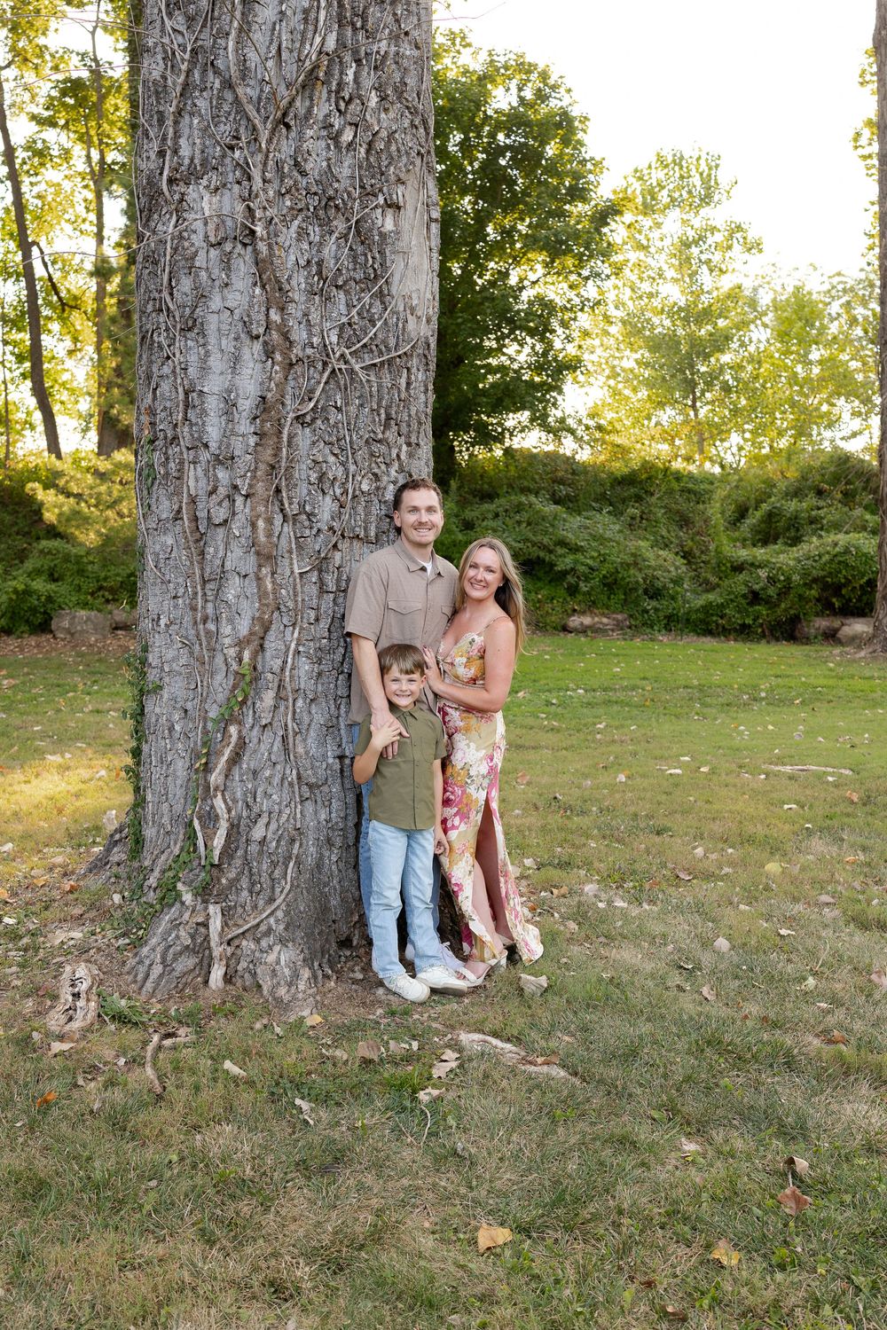 Family of three poses together against a large tree trunk in a sunny park with green trees and fallen leaves on the grass.