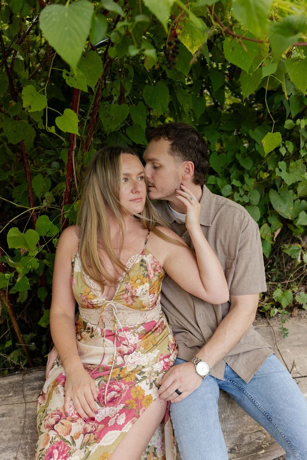 Couple sitting close on a wooden bench in front of green leafy vines, the man kissing the woman's temple as she wears a floral sundress.